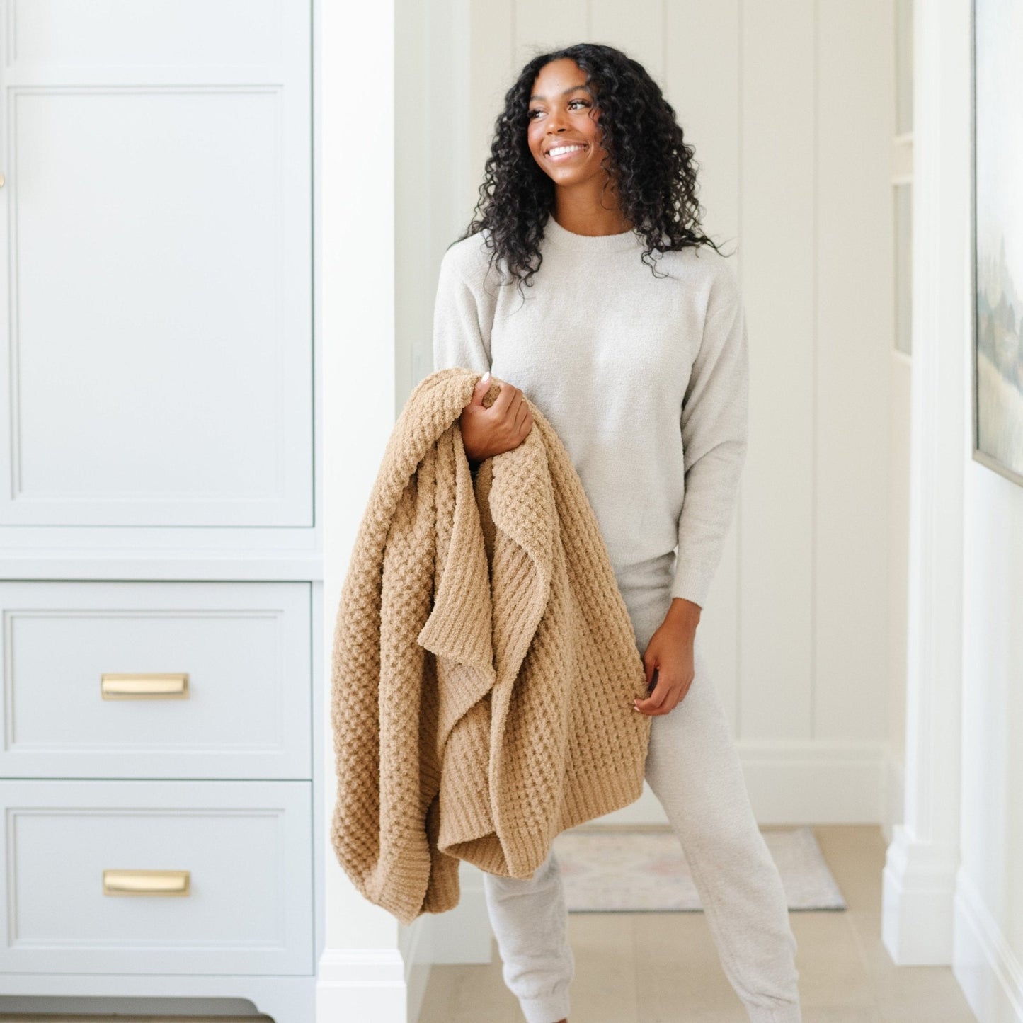 Person holding a folded, tan waffle knit throw blanket at home..
