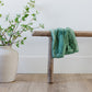 A Mini Seagrass (dusty green) Colored Lush Saranoni Blanket lays over a wood bench. The soft blanket is a small blanket and a baby blanket or toddler blanket.