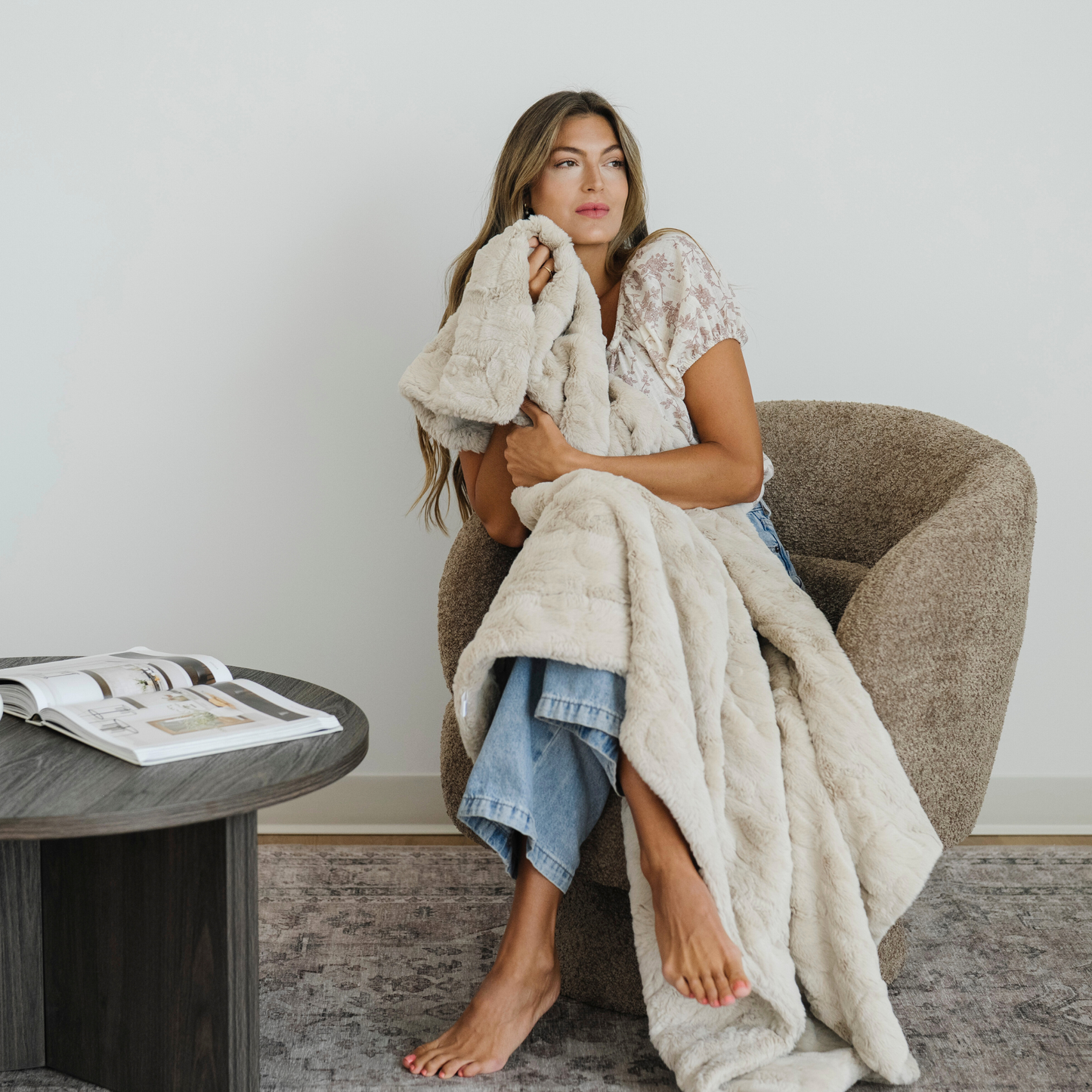 Woman sitting in chair snuggled with a tan/cream faux fur soft cozy throw blanket. 