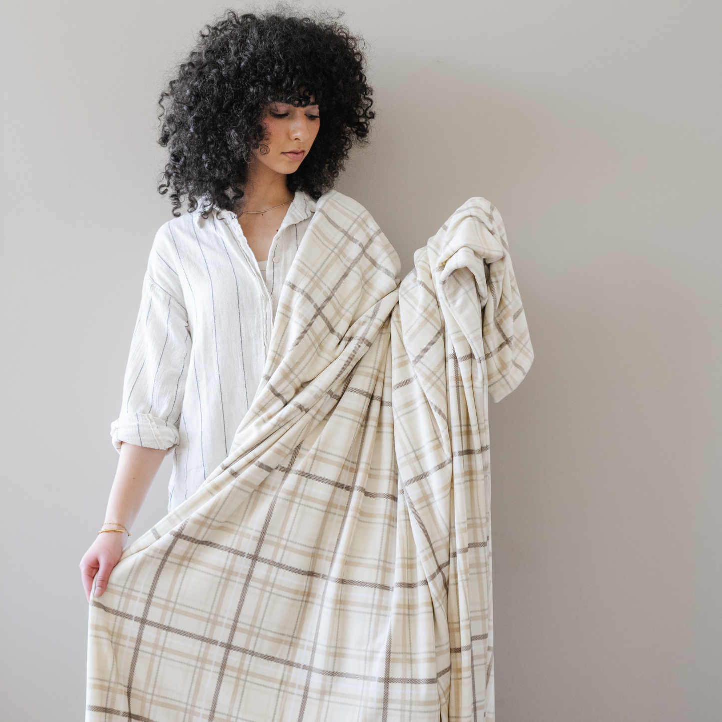 Woman stands snuggled with a plaid minky stretch throw blanket. 