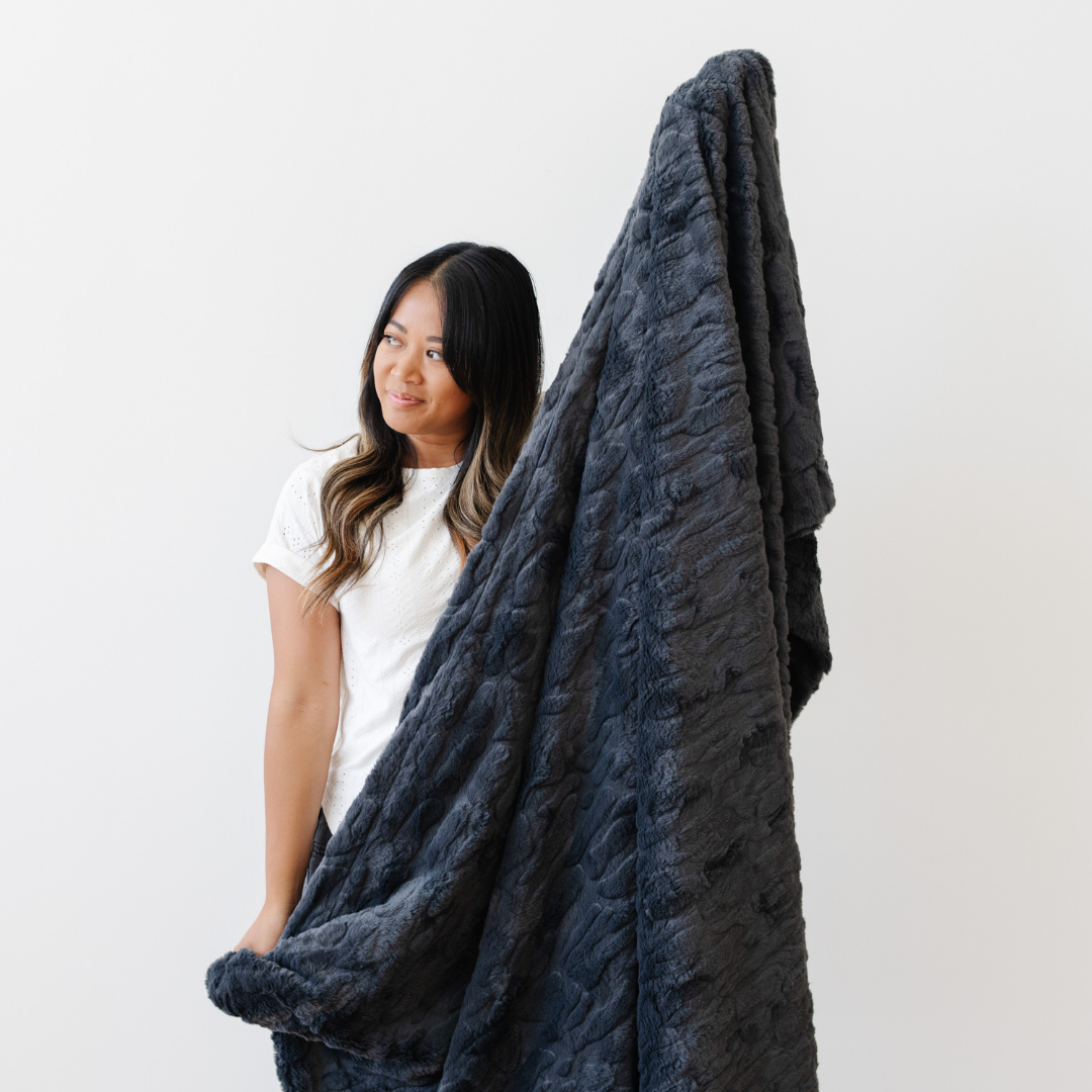 A woman smiles and holds a charcoal luxurious patterned faux fur extra large throw blanket that is a Saranoni blanket. This ultra-soft, cozy blanket adds a touch of elegance and warmth to any space, perfect as a soft oversized blanket or fluffy blanket for lounging. Ideal for those seeking a high-quality artificial fur blanket or luxury blanket, this versatile and stylish soft blanket enhances both comfort and decor. Shop the best blankets and throws for ultimate relaxation and cozy vibes.