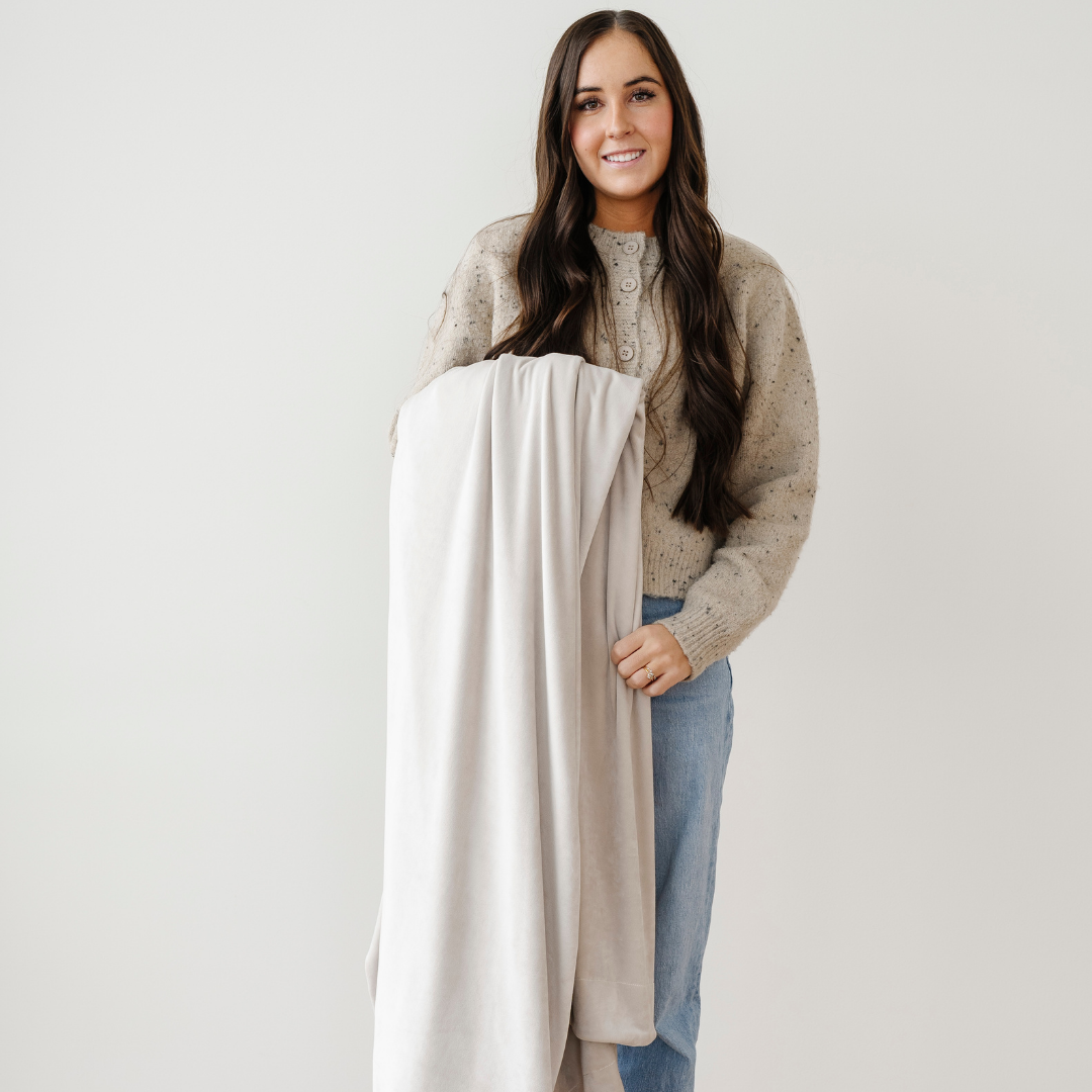 A woman is holding a light neutral minky stretch blanket. This Saranoni blanket is a throw blanket and is super stretchy and durable. Enjoy this cozy blanket for years to come. 