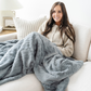 A woman sits on a couch with a blue patterned faux fur blanket. This soft blanket has a luxurious texture and is a Saranoni blanket. This cozy blanket is a great comfort gift, loungewear blanket, or home decor staple. 