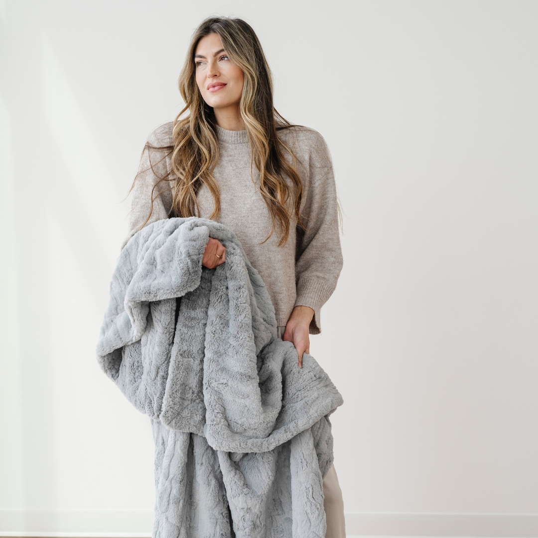 A woman holds a gray blanket that is a patterned faux fur blanket. This blanket has a luxurious texture and is a Saranoni blanket. This luxury blanket can be used for home decor, relaxing, or gifting.  