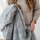 A luxurious gray patterned faux fur blanket that is being held by a woman. This is a Saranoni blanket and also very durable. This soft blanket can be used for gifting, relaxing, or home decor. 