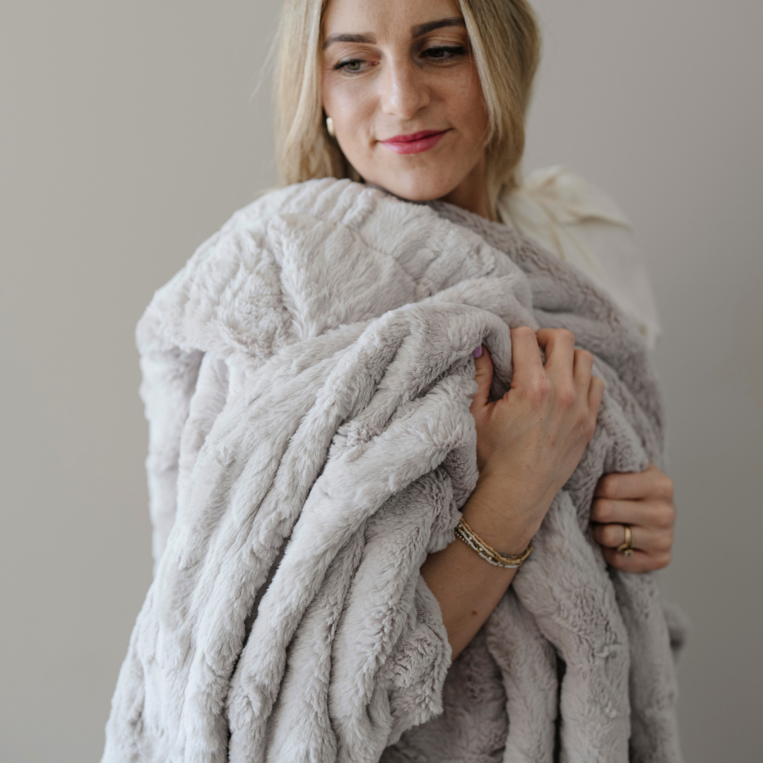A woman holds a light gray patterned faux fur blanket that is a Saranoni blanket. This throw blanket is luxuriously soft and is great for gifting, relaxing, and using for home decor. This blanket is also very durable and cozy. 