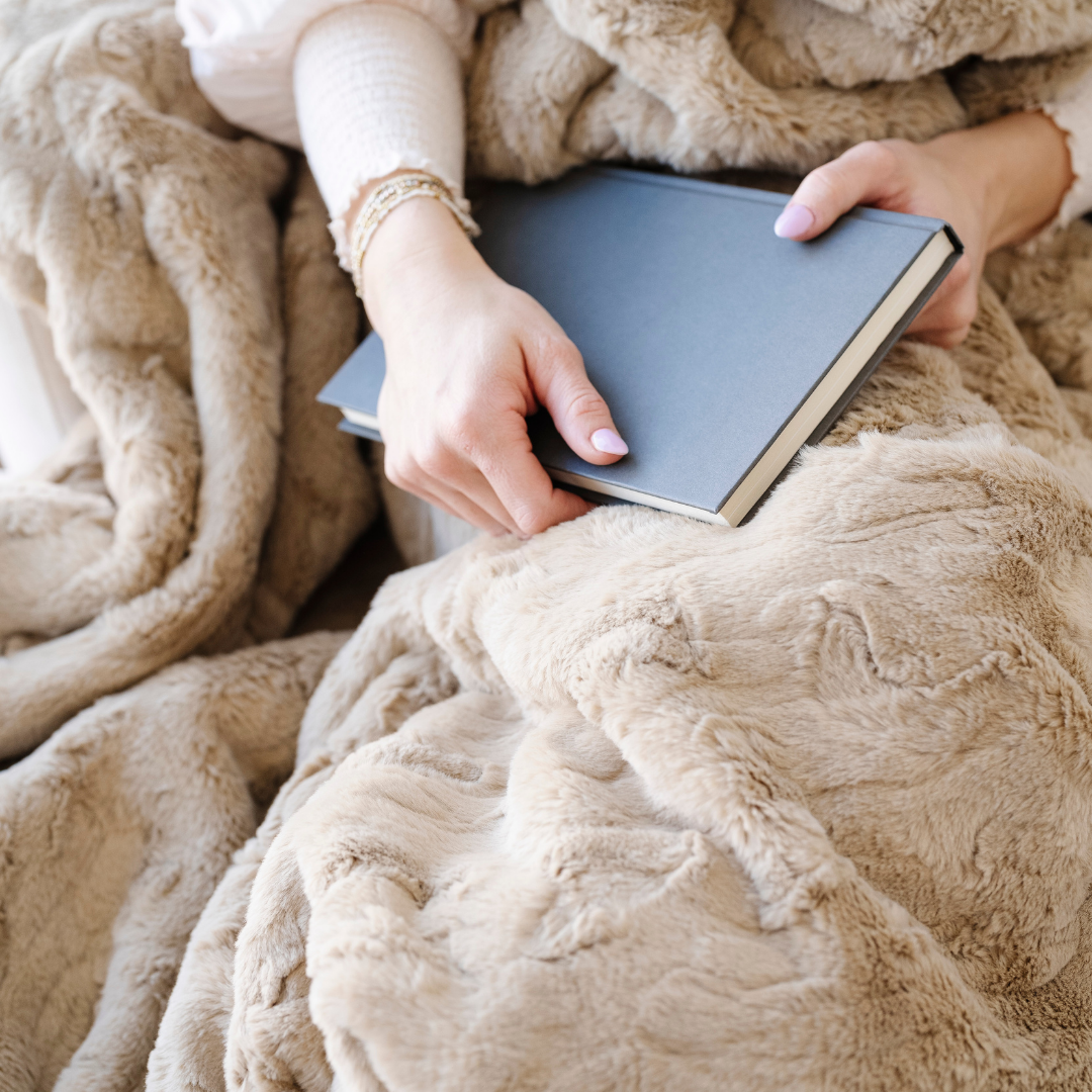 A woman is holding a book and has a khaki patterned faux fur blanket on her lap. This Saranoni blanket is a luxury blanket and has an incredibly soft texture. Elevate your luxury living room by adding this blanket to your home. 