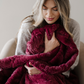 A woman holds a deep red patterned faux fur blanket. This soft blanket is a Saranoni blanket that has a very luxurious texture. Give this blanket as a comfort blanket or add to your home decor staples. 
