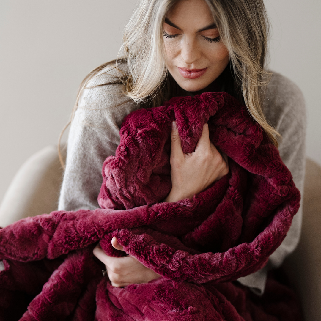 A woman holds a deep red patterned faux fur blanket. This soft blanket is a Saranoni blanket that has a very luxurious texture. Give this blanket as a comfort blanket or add to your home decor staples. 