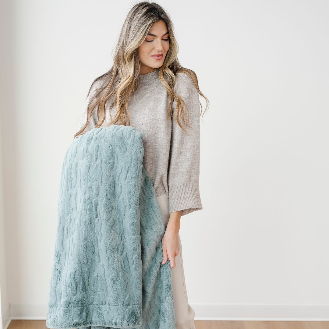 A woman holds a eucalyptus colored patterned faux fur blanket. This Saranoni blanket has a very luxurious texture. Experience luxury living with this throw blanket. 