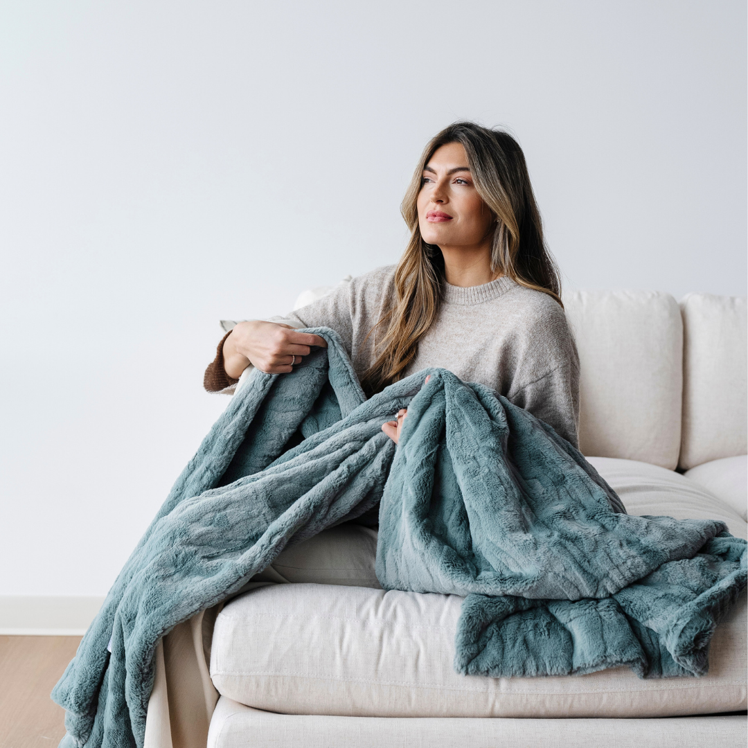 A woman sits on a couch with with a eucalyptus patterned faux fur blanket. This throw blanket has a luxurious texture and is perfect for luxury living. 