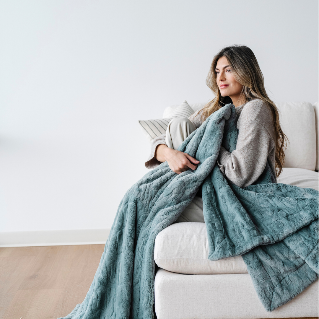 A woman sits on a couch and she is holding a eucalyptus patterned faux fur blanket. Add this blanket to a luxury living room or cozy house. This throw blanket is also great for relaxing or gifting. Enjoy this Saranoni luxury blanket for years to come. 