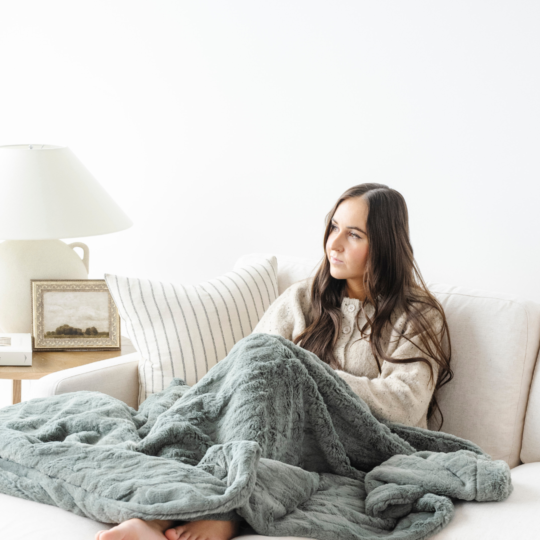 A woman sits on a couch of with a green patterned faux fur blanket. This soft blanket is a Saranoni blanket and has a luxurious texture. This cozy blanket is great for gifting, lounging, or home decor. 