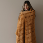 A woman is wrapped in a patterned faux fur blanket that is a copper color. This Saranoni blanket is a very durable and soft blanket. This luxury blanket is perfect for gifting, lounging, and as home decor. 