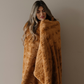 A woman is wrapped in a Saranoni blanket that is a copper colored patterned faux fur blanket. This throw blanket is perfect for home decor, adding to a luxury living room, or refreshing a bedroom. Enjoy this soft blanket for years to come. 