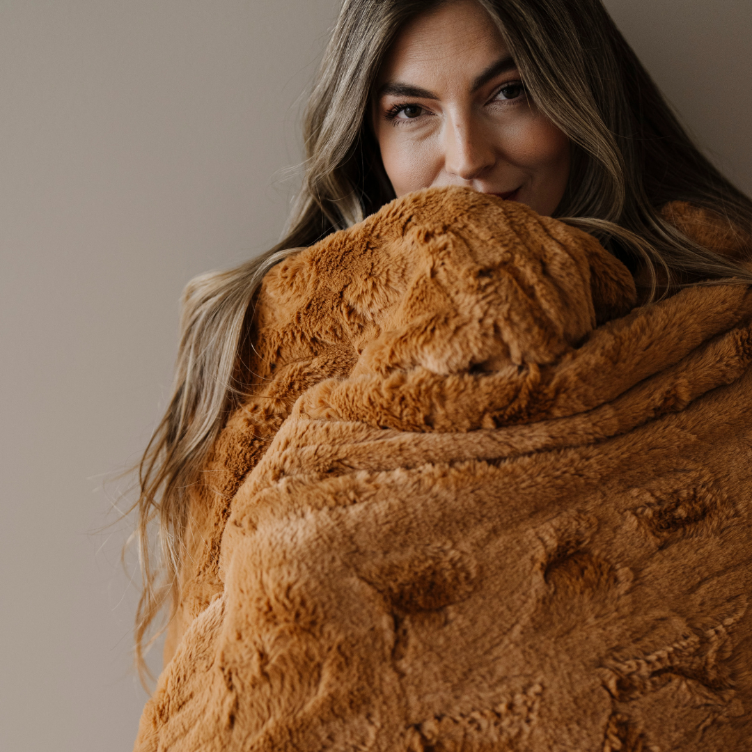 A woman is wrapped in a copper colored patterned faux fur blanket. This Saranoni luxury blanket has a super plush texture. This throw blanket is perfect for gifting, relaxing, or home decor. 