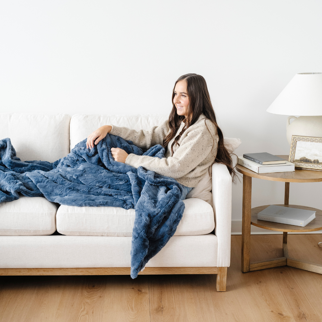 A woman is sitting on a cream couch and has a blue patterned faux fur blanket on her lap. This is a Saranoni blanket that has a super plush texture and is great for cozy home decor. This throw blanket is also perfect for relaxing and gifting. 