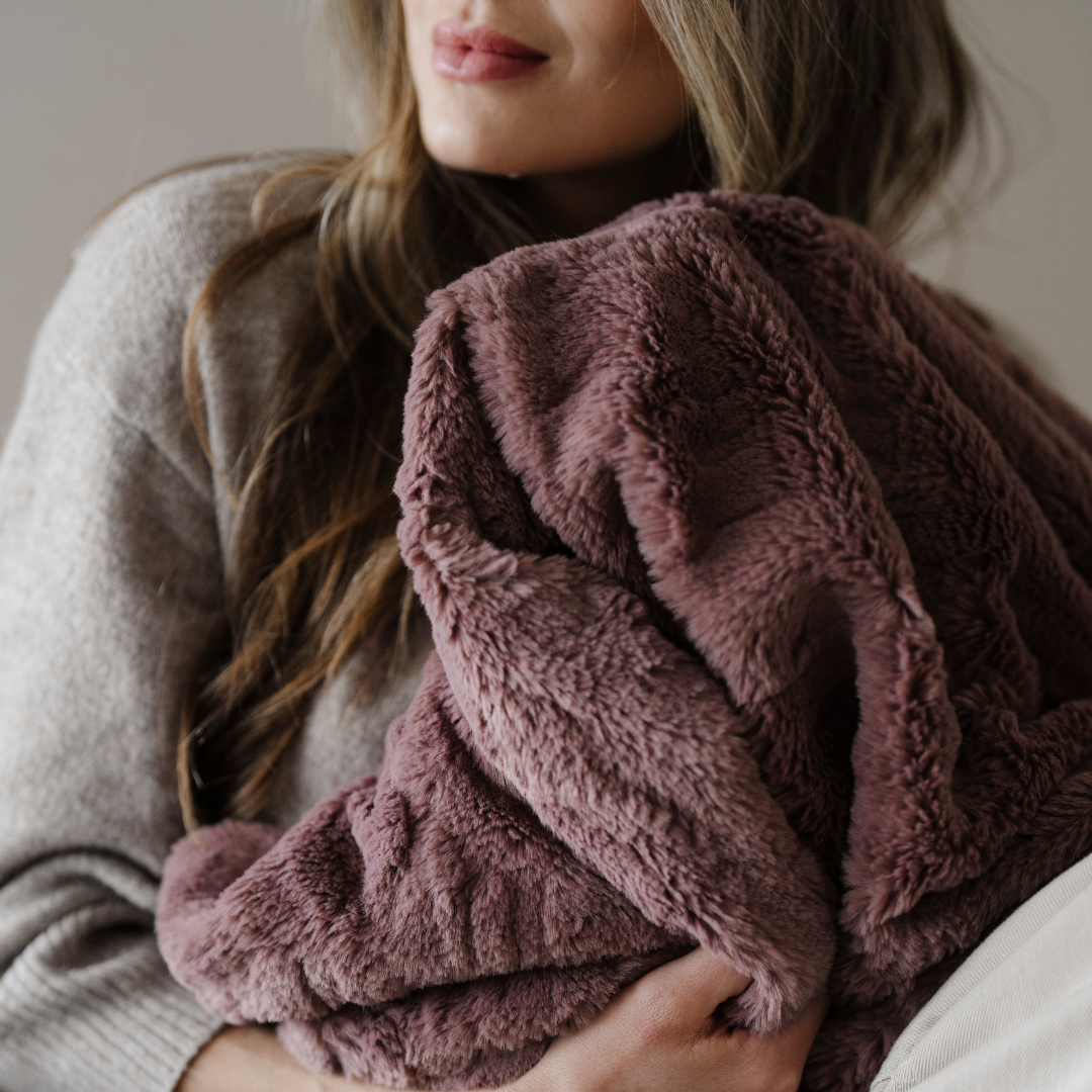 A woman is holding a dusty mauve patterned faux fur blanket. This soft blanket is a Saranoni blanket. Use this luxury blanket as a comfort gift, for lounging, or home decor. 
