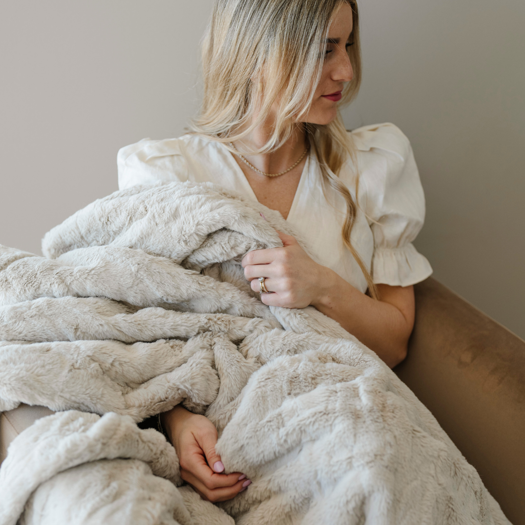 A woman holds a light tan patterned faux fur blanket. This soft blanket is a Saranoni blanket. This luxury throw is perfect for gifting, lounging or home decor. It's luxurious texture makes it an extra cozy blanket. 