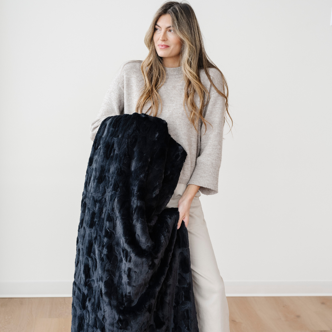 A woman holds a black patterned faux fur blanket. This luxury blanket is from Saranoni and has a plush texture. Add this throw blanket to any bedroom or living room to make the space cozier. 