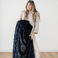 A woman holds a black patterned faux fur blanket. This luxury blanket is from Saranoni and has a plush texture. Add this throw blanket to any bedroom or living room to make the space cozier. 