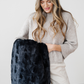 A woman is looking at a black patterned faux fur blanket while holding it. This soft blanket is a Saranoni blanket. This blanket is perfect for gifting, lounging, and home decor. 