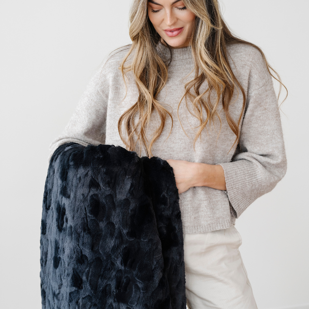 A woman is looking at a black patterned faux fur blanket while holding it. This soft blanket is a Saranoni blanket. This blanket is perfect for gifting, lounging, and home decor. 