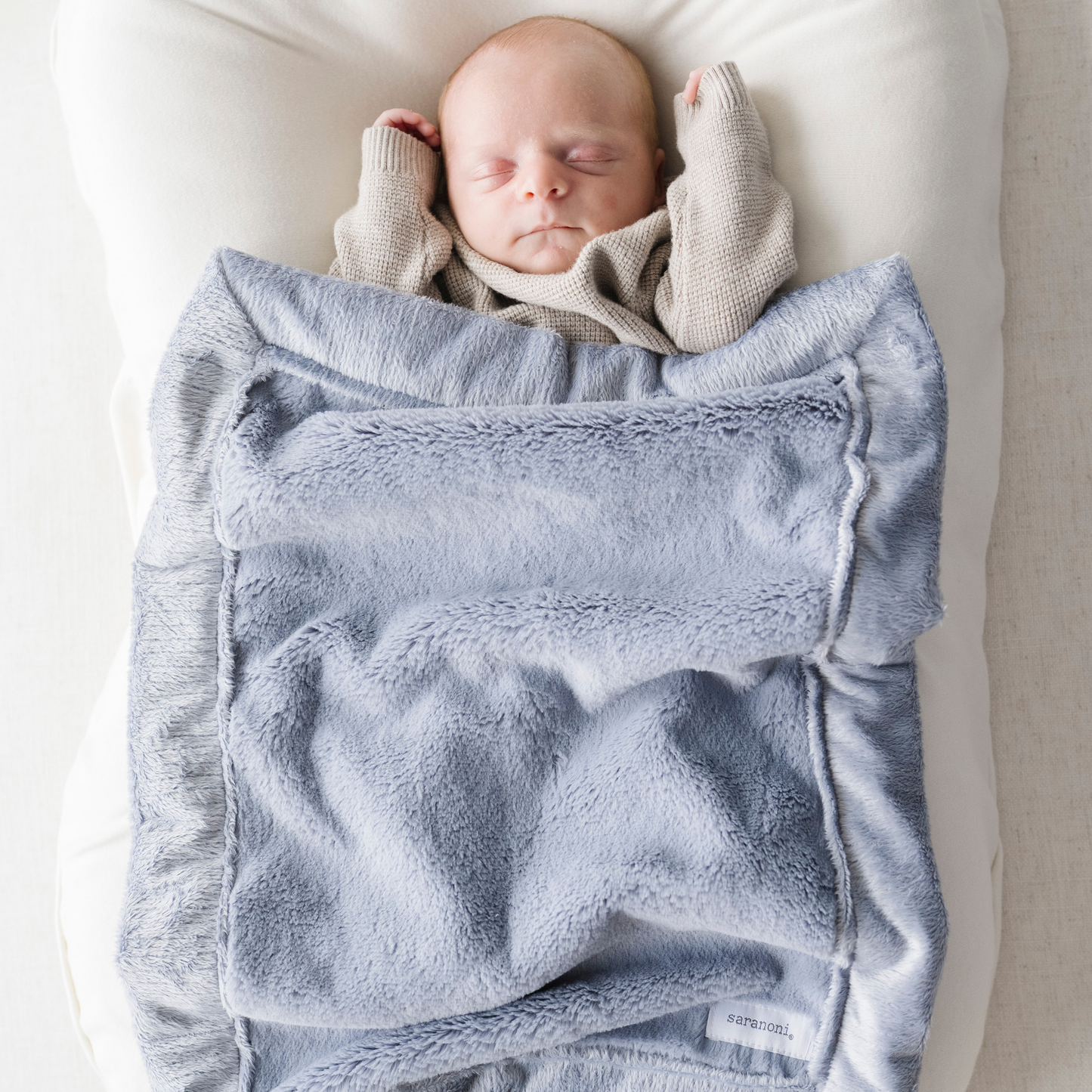 A baby boy is laying under a blue mini blanket from Saranoni.