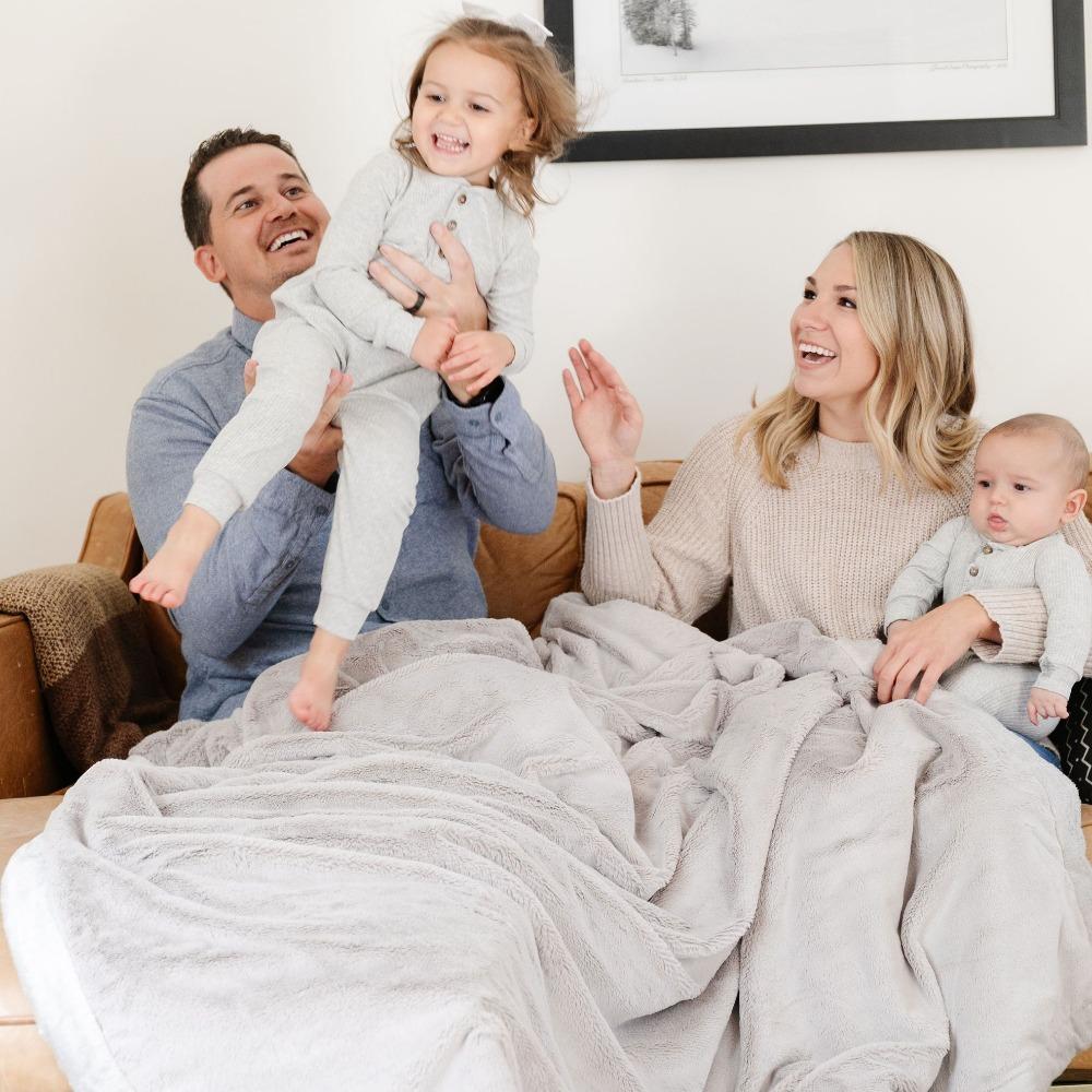 A husband and wife and their son and daughter play together on a Luxury Extra Large Oversized Throw Feather (light gray) Colored Lush Saranoni Blanket. The soft blanket is an extra large throw blanket and has a soft faux fur feeling.