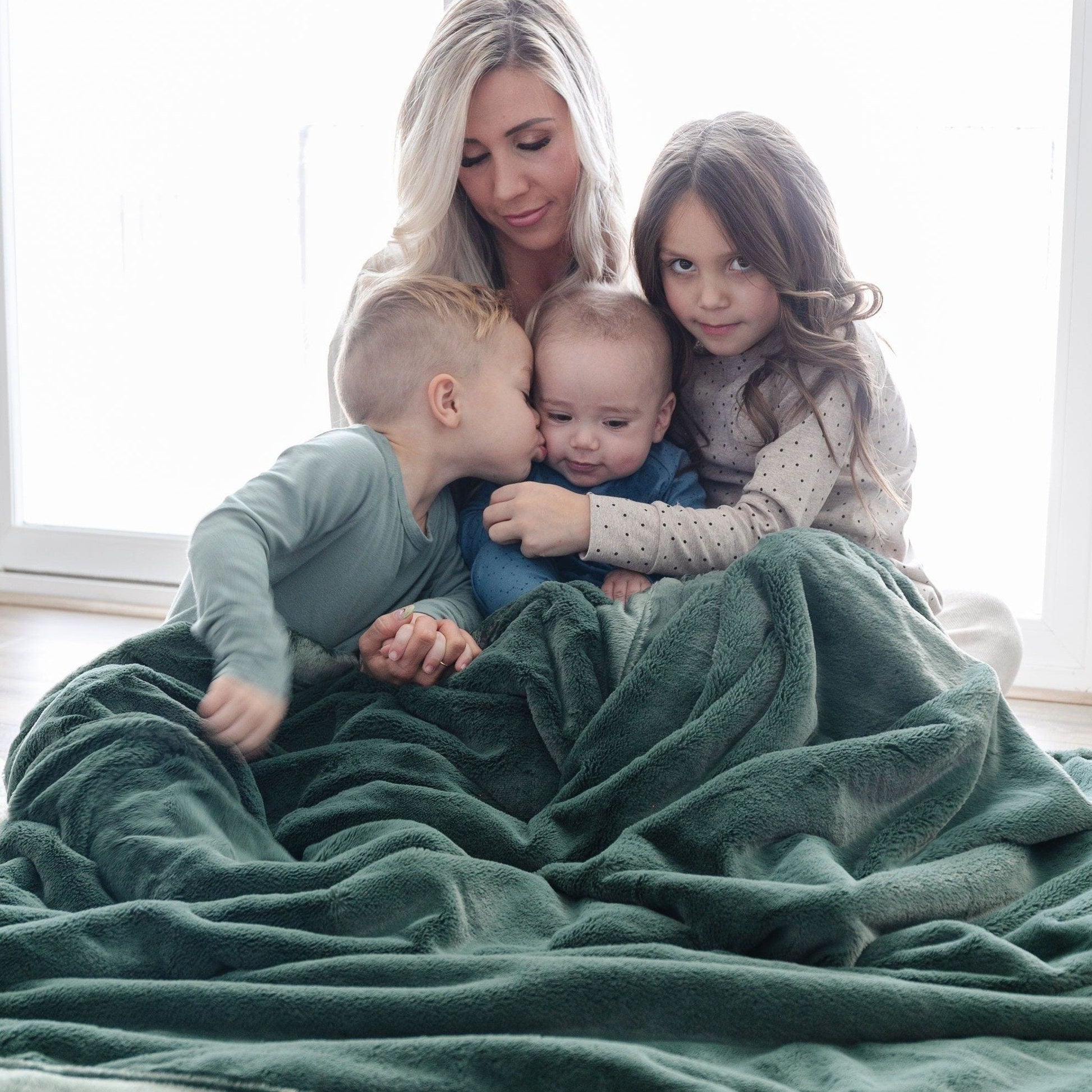 A mother and her two sons and daughter sit under a Luxury Extra Large Oversized Throw Hunter Green Colored Lush Saranoni Blanket. The soft blanket is an extra large throw blanket and has a soft faux fur feeling.