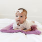 A little baby girl with a white bow and a white outfit lays on a Mini Saranoni Lush Blanket that is a lighter purple color. The specific color is called Fairy Wings.