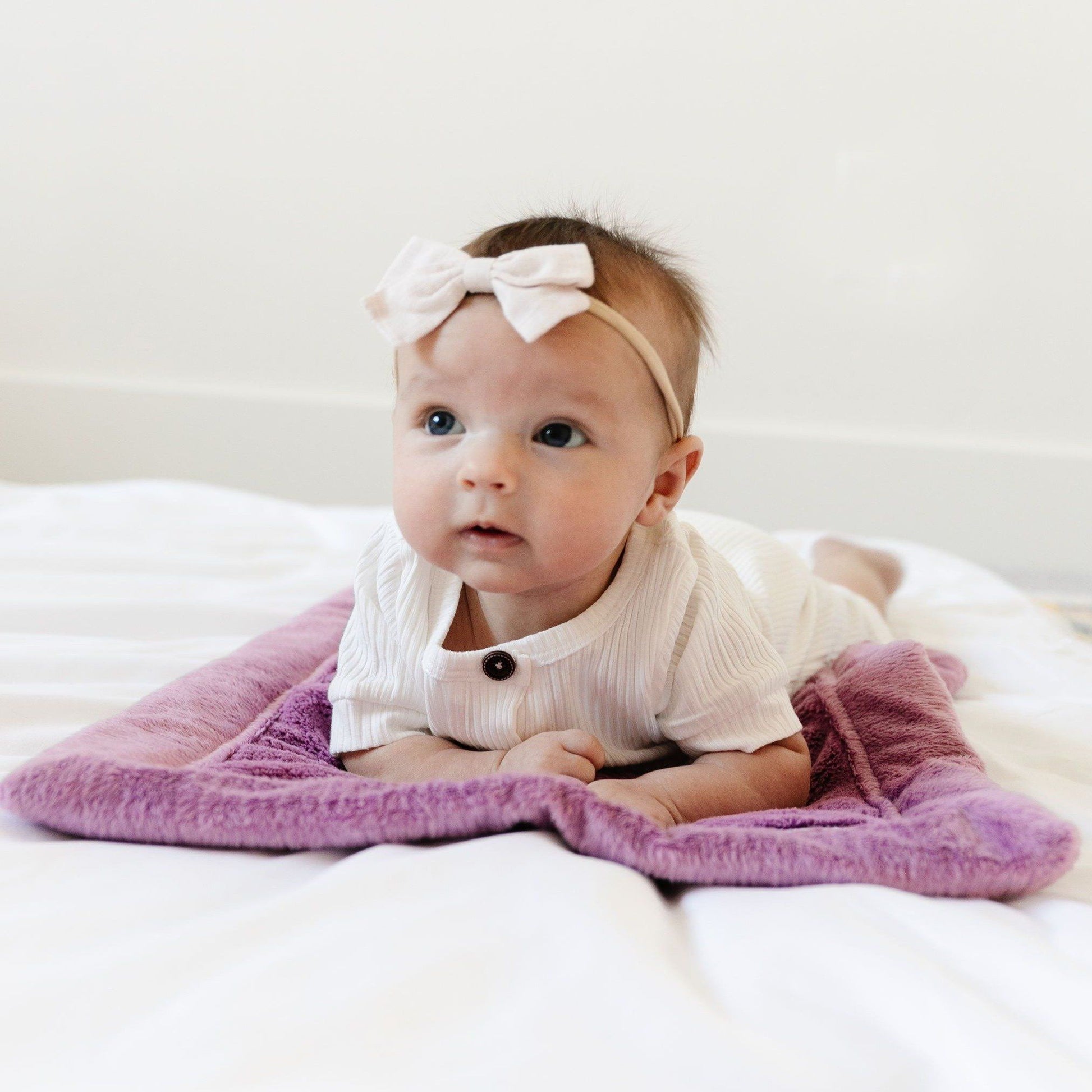 A little baby girl with a white bow and a white outfit lays on a Mini Saranoni Lush Blanket that is a lighter purple color. The specific color is called Fairy Wings.