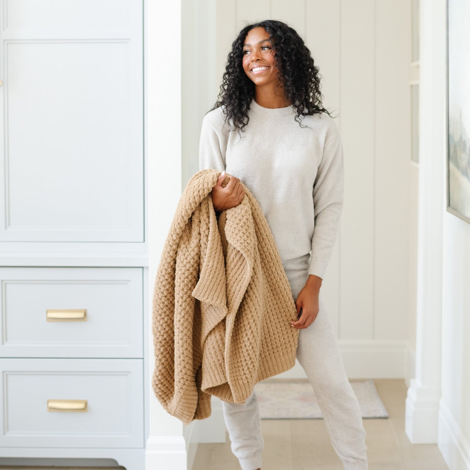 Person holding a folded, tan waffle knit throw blanket at home..