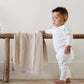 A baby boy wearing a white Saranoni Cotton Set stands by a soft, tan Saranoni Heirloom Cotton Knit Receiving Blanket. The luxury 100% cotton blanket features classic cable knit detailing, a wide border, making it a timeless keepsake. The cozy, elevated blanket, is perfect for special occasions like baby showers or newborn photoshoots. Ideal for searches related to personalized baby blankets, soft cotton receiving blankets, or heirloom-quality baby gifts.
