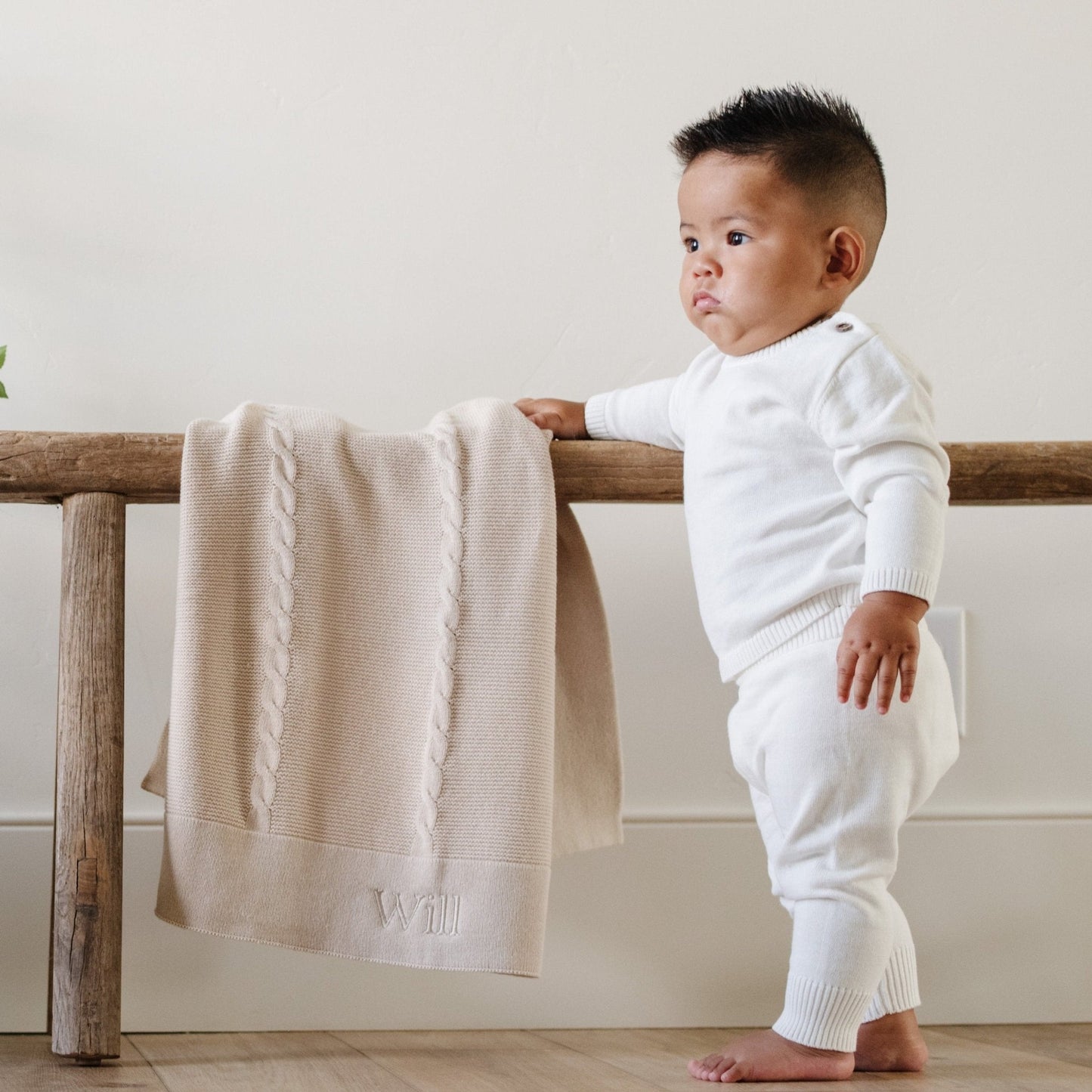A baby boy wearing a white Saranoni Cotton Set stands by a soft, tan Saranoni Heirloom Cotton Knit Receiving Blanket. The luxury 100% cotton blanket features classic cable knit detailing, a wide border, making it a timeless keepsake. The cozy, elevated blanket, is perfect for special occasions like baby showers or newborn photoshoots. Ideal for searches related to personalized baby blankets, soft cotton receiving blankets, or heirloom-quality baby gifts.