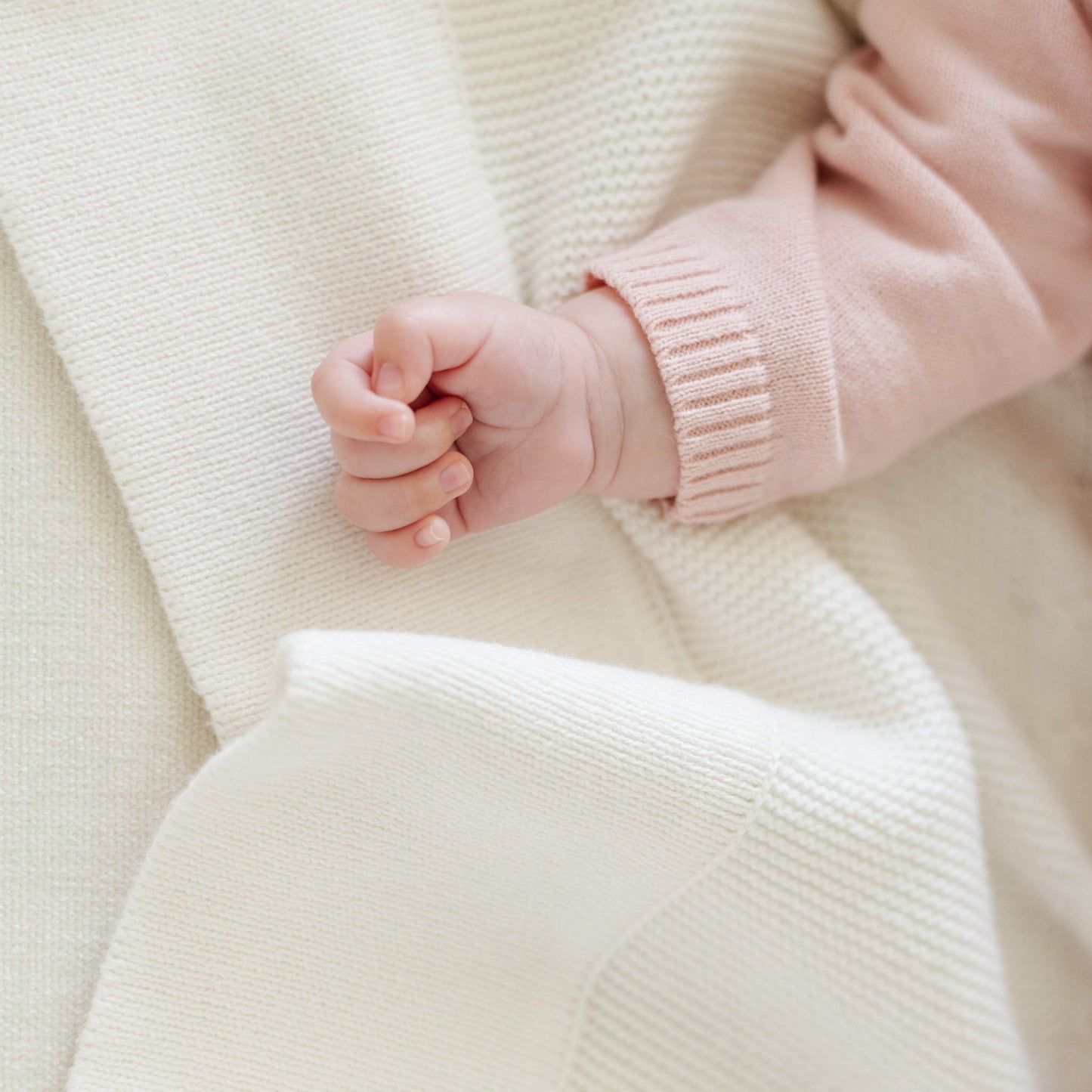 A baby girl lays on a soft, white Saranoni Heirloom Cotton Knit Receiving Blanket. The luxury 100% cotton blanket features classic cable knit detailing, a wide border, making it a timeless keepsake. The cozy, elevated blanket, is perfect for special occasions like baby showers or newborn photoshoots. Ideal for searches related to personalized baby blankets, soft cotton receiving blankets, or heirloom-quality baby gifts.