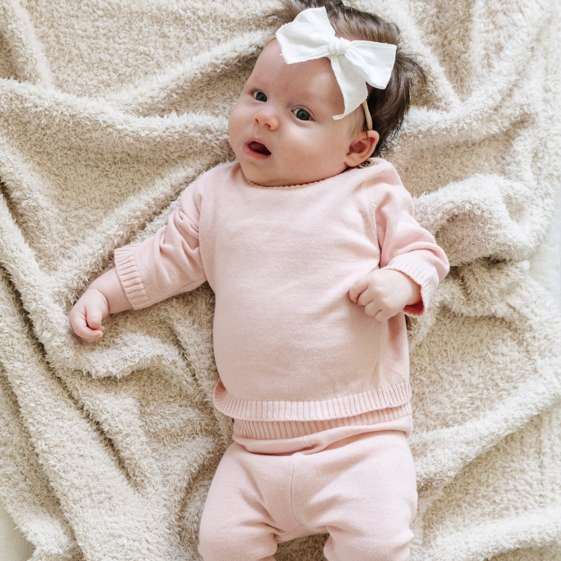 A baby girl lays on a Saranoni Blanket. She is wearing a light pink, 100% cotton Saranoni baby outfit that combines style and comfort. The Saranoni cloud-soft cotton set features a breathable waistband, loosely fitted sleeves, and elevated button details, making it perfect for everyday wear or special occasions. This heirloom-quality baby clothing offers timeless style and ultimate comfort. Ideal for parents searching for luxury baby clothes, soft cotton baby sets, or high-quality infant outfit.